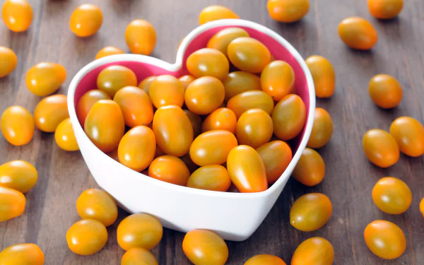 Heart-shaped white bowl filled with small orange tomatoes on a wooden surface, captured in vibrant 4K Ultra HD clarity as a desktop wallpaper background.