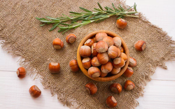 A rustic bowl filled with hazelnuts sits on a textured burlap background, accompanied by fresh rosemary, showcasing food in stunning 8K Ultra HD detail.