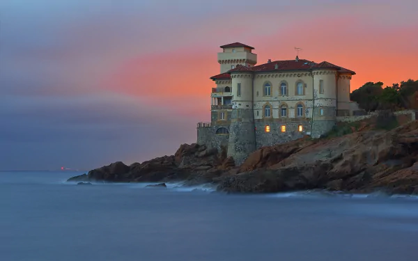 HD wallpaper of a historic lighthouse building on the rocky coast of Livorno, Italy, overlooking the calm ocean at sunset with a colorful sky.