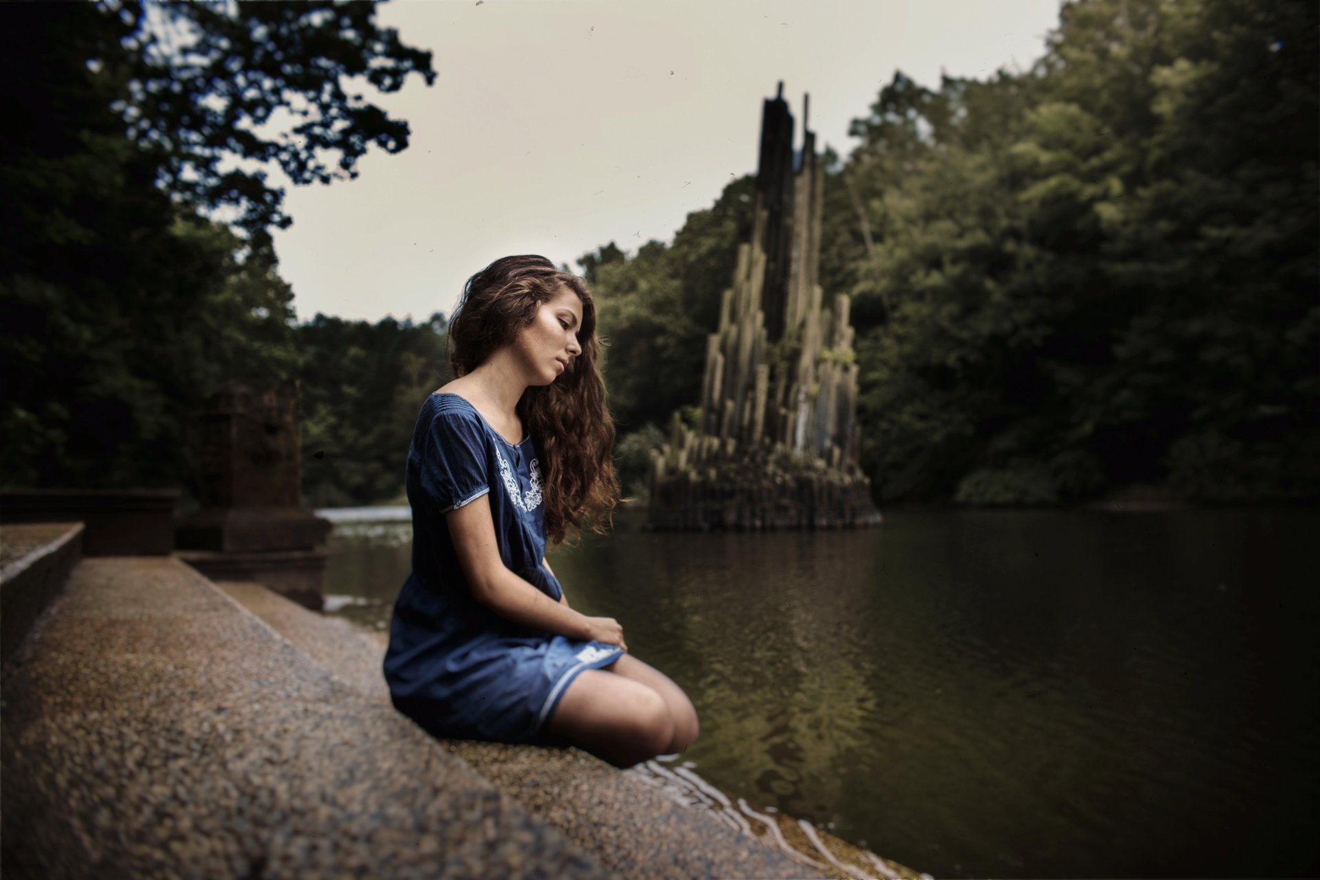 Brunette woman model in a contemplative mood sits by a serene outdoor lake, surrounded by lush greenery; captured in 4K Ultra HD for a vivid desktop wallpaper.