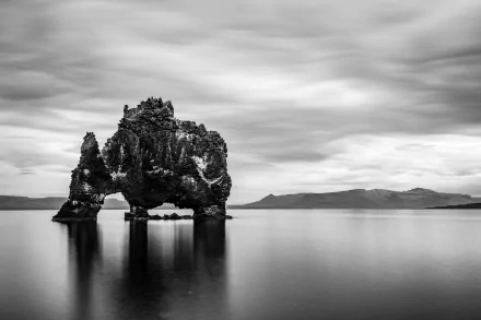 Black and white HD desktop wallpaper featuring a striking natural rock arch rising from calm waters under a cloudy sky, blending serene nature with rugged texture.