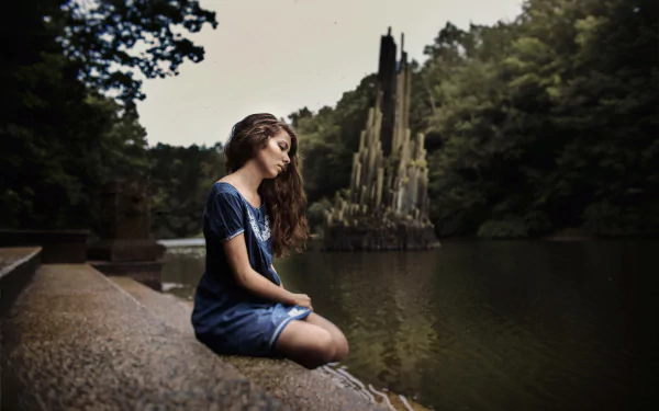 Brunette woman model in a contemplative mood sits by a serene outdoor lake, surrounded by lush greenery; captured in 4K Ultra HD for a vivid desktop wallpaper.