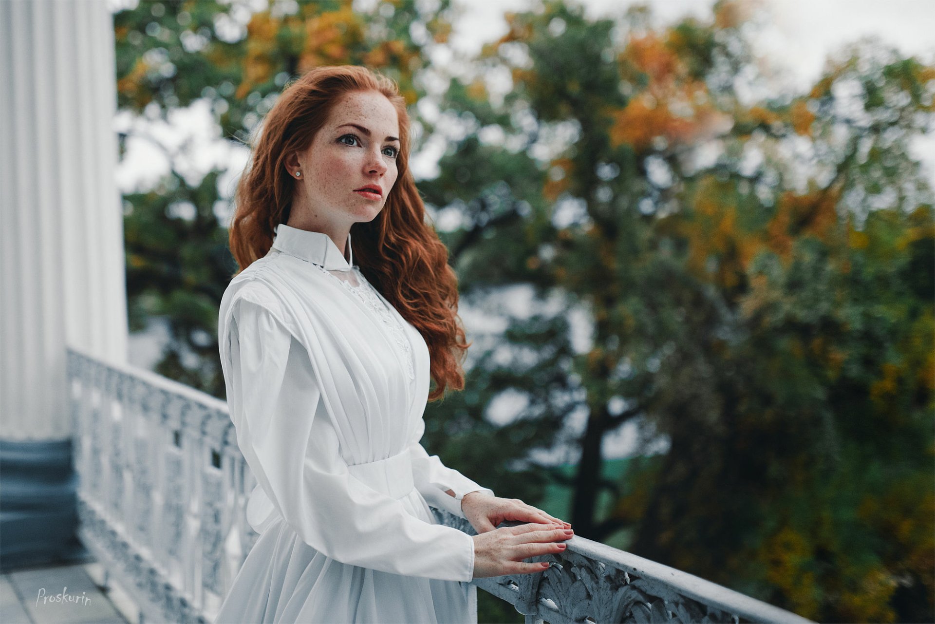 Redhead woman with freckles in a white dress stands on a porch railing, surrounded by autumn foliage and soft bokeh, captured in an HD desktop wallpaper.