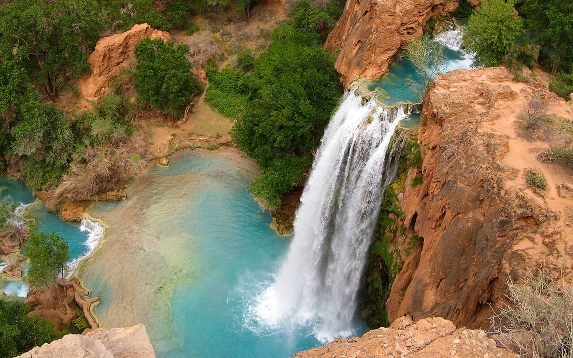 HD PC desktop wallpaper: aerial view of Havasu Falls — turquoise pool and white cascade over red rock cliffs, surrounded by green trees; vivid nature waterfall background.