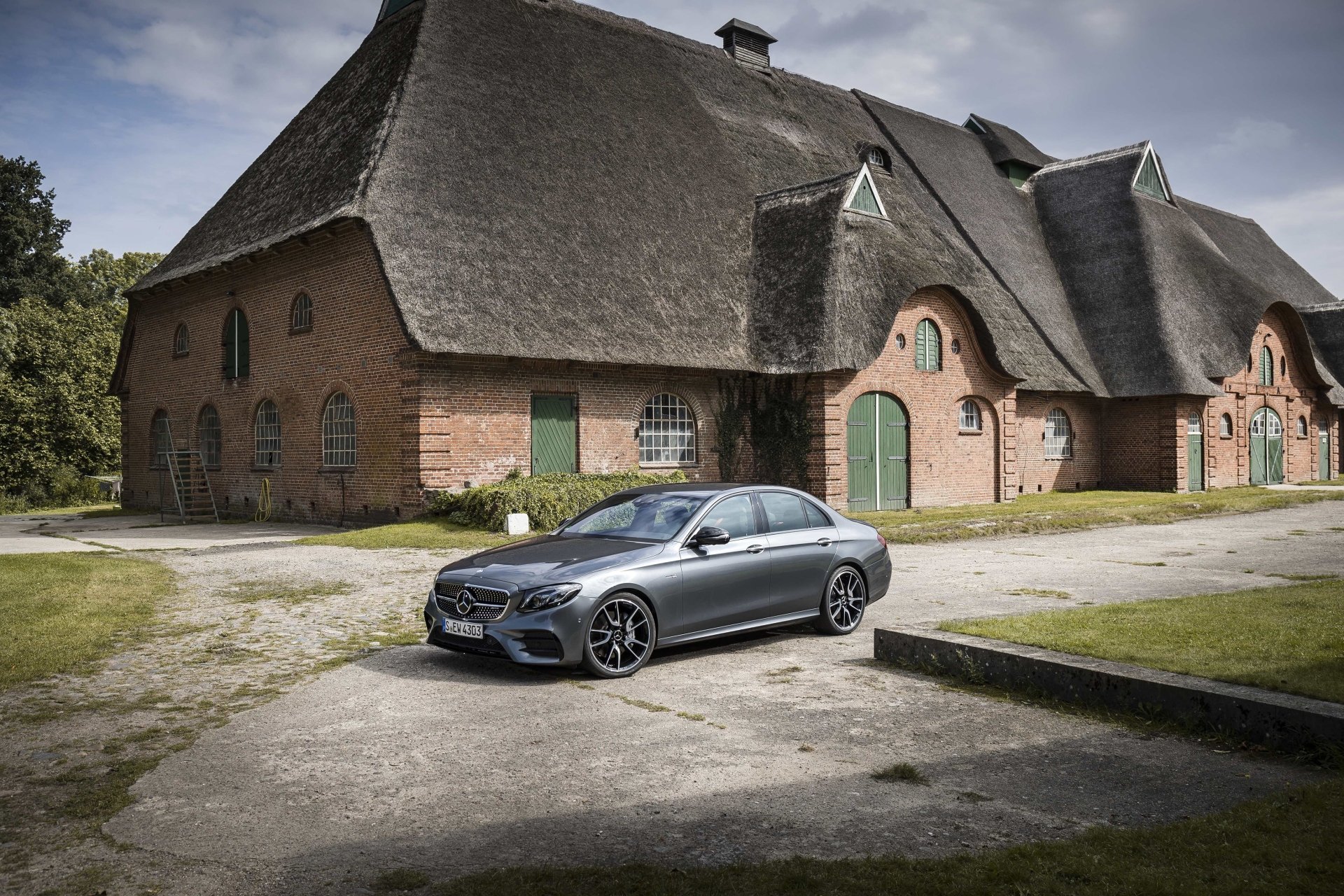 A silver Mercedes-Benz E-Class sedan parked on a driveway in front of a traditional thatched-roof building, captured in 4K Ultra HD for a desktop wallpaper.