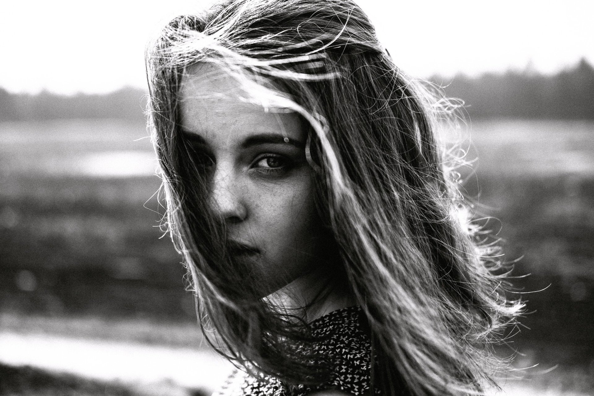 Black and white HD desktop wallpaper of a woman’s face in sunlight, with wind-swept hair and a blurred natural background.