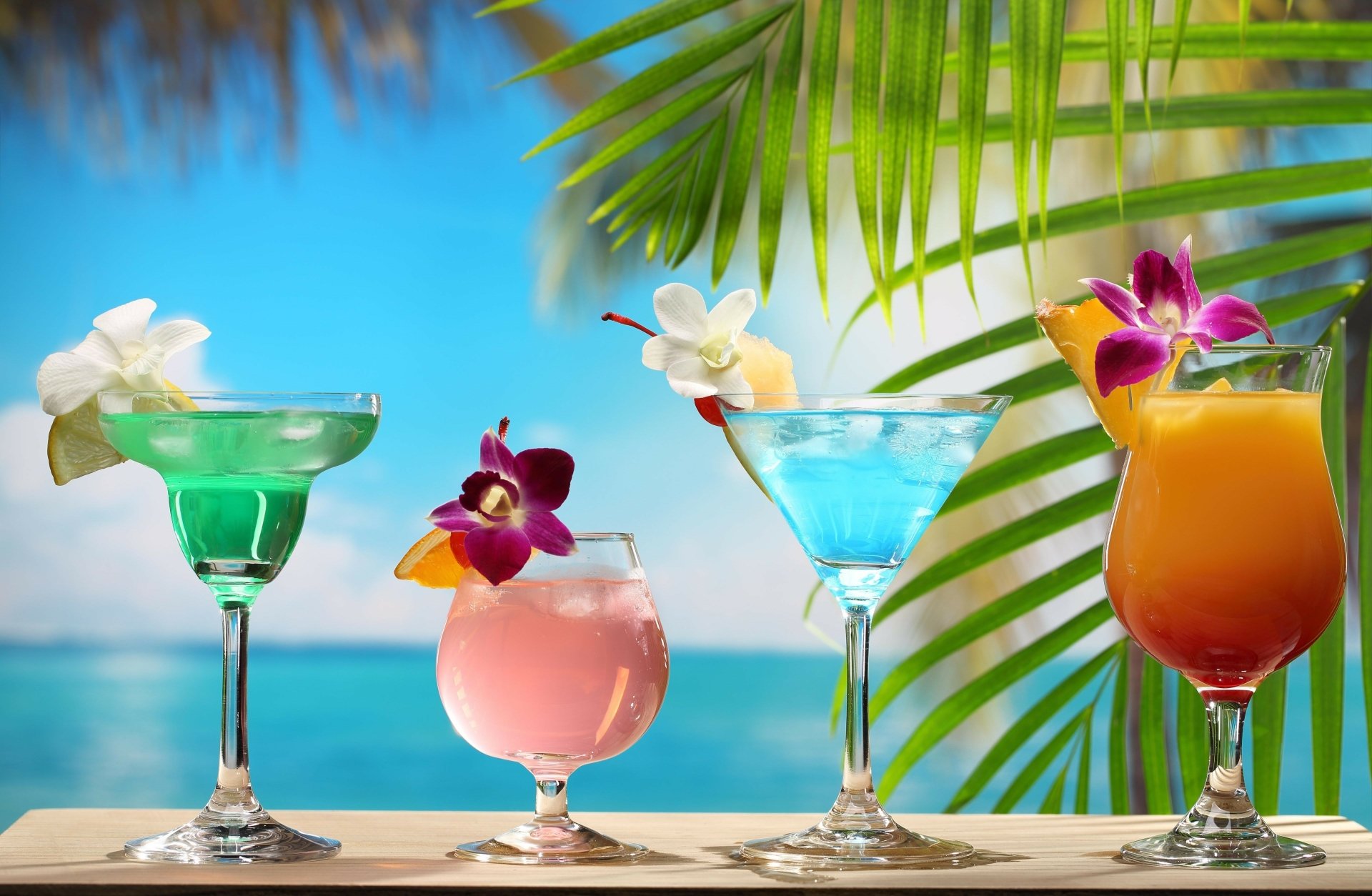 A vibrant 4K Ultra HD wallpaper featuring colorful summer cocktails in glassware, decorated with flowers, set against a tropical beach backdrop with palm leaves.