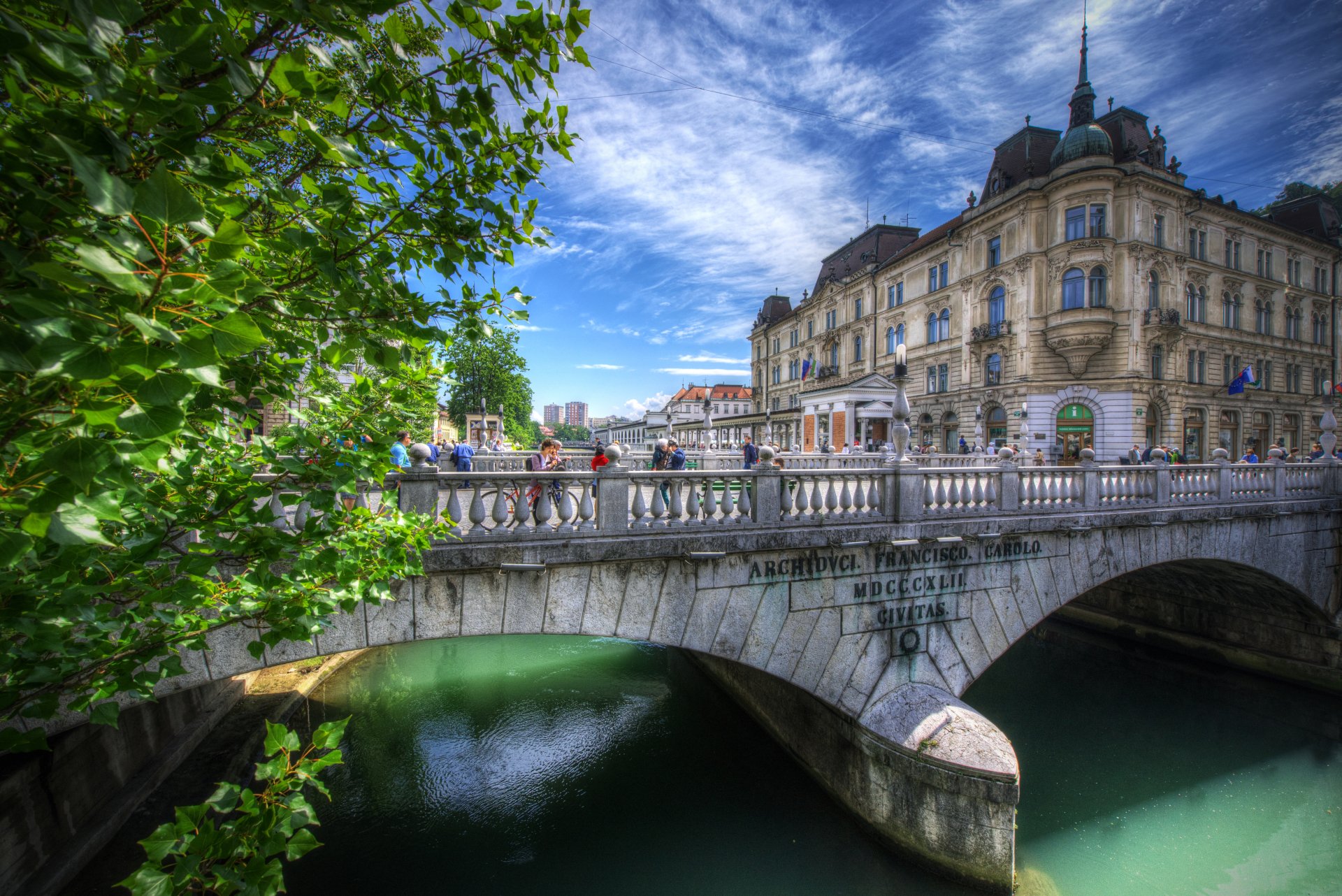 HDR 5K Ultra HD desktop wallpaper of Ljubljana, Slovenia: ornate city buildings and a stone bridge over a canal, framed by leafy trees under a dramatic sky.