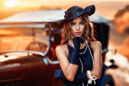 A brunette woman with brown eyes, wearing a hat, gloves, and pearl accessories, stands in front of a vintage car at sunset. This HD desktop wallpaper features the model in elegant attire.