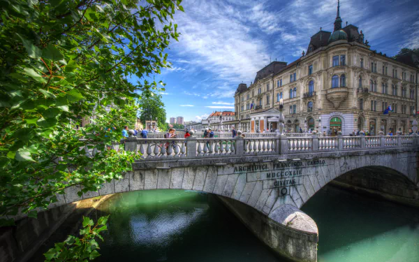 HDR 5K Ultra HD desktop wallpaper of Ljubljana, Slovenia: ornate city buildings and a stone bridge over a canal, framed by leafy trees under a dramatic sky.