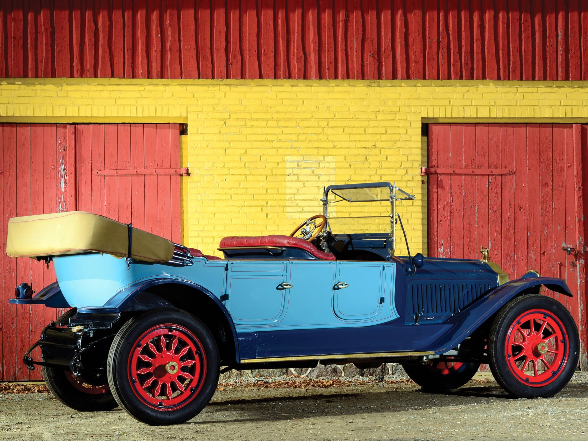 HD PC wallpaper featuring a vintage 1914 Packard Six 7-passenger Touring car, painted blue with red wheels, parked in front of a yellow brick wall and red doors.
