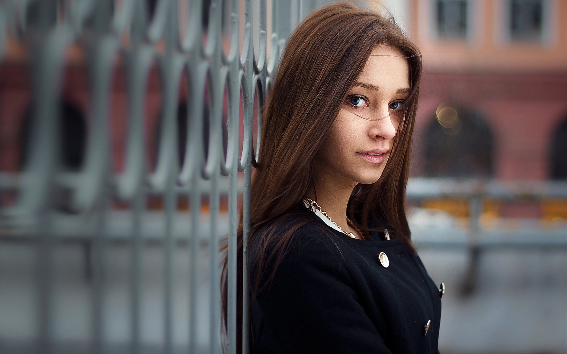 A brunette woman with blue eyes leans against a metal fence, wearing a black coat. The background shows urban buildings in soft focus. HD desktop wallpaper and background.
