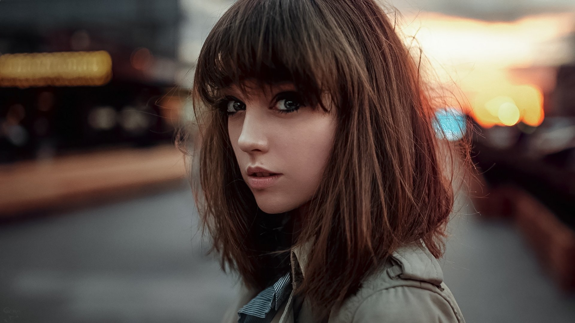 Brunette model with striking features gazes intently at the camera, set against a soft, blurred urban background. High-definition wallpaper showcasing beauty and depth.