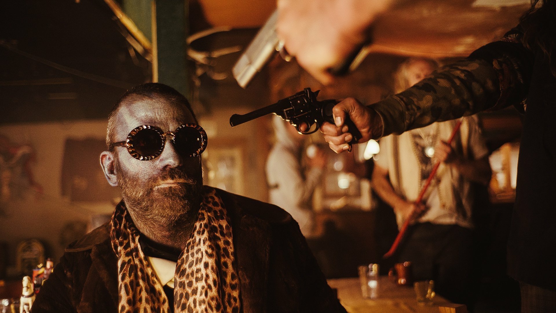 A dramatic scene from the TV show Z Nation, featuring a man with sunglasses and a leopard-print scarf, confronted by a gunman in a dimly lit bar. HD desktop wallpaper.