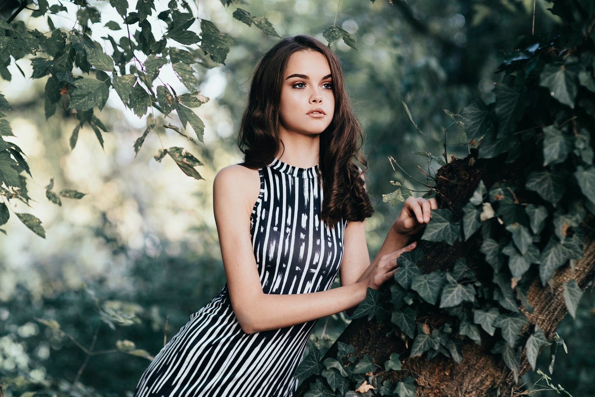 Brunette woman in a striped dress stands confidently in a lush outdoor setting, leaning against a tree, with ivy and foliage in the background. HD desktop wallpaper and background.