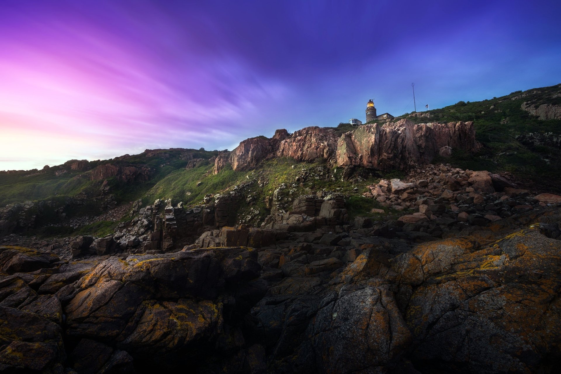 HD PC desktop wallpaper and background: man-made lighthouse atop rugged coastal cliffs at dusk, vivid purple and blue sky over a rocky foreground.