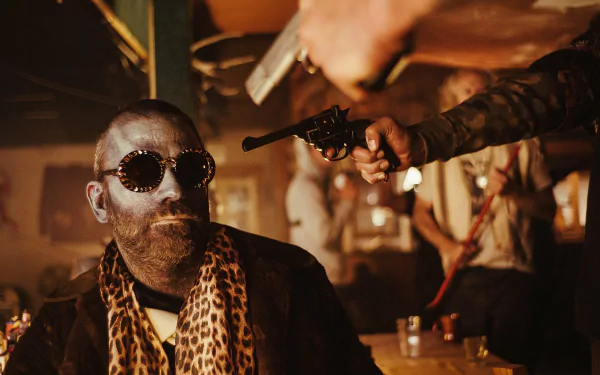 A dramatic scene from the TV show Z Nation, featuring a man with sunglasses and a leopard-print scarf, confronted by a gunman in a dimly lit bar. HD desktop wallpaper.