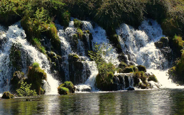 HD PC desktop wallpaper: nature waterfall cascading over mossy tiers into a calm reflective pool, framed by lush green foliage.