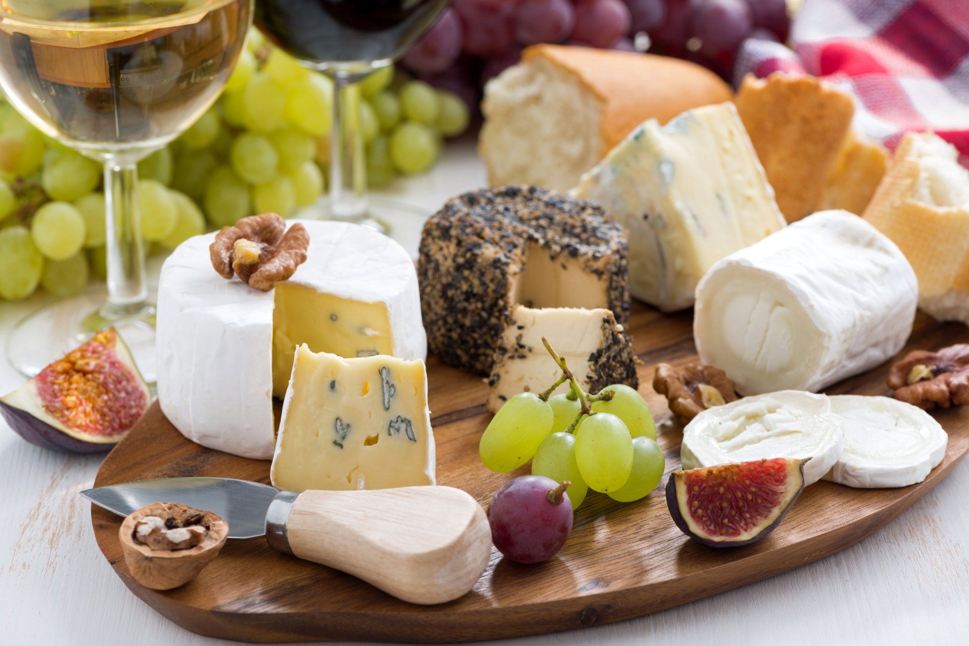 A 4K Ultra HD still life PC wallpaper featuring an assortment of cheeses, grapes, figs, walnuts, and glasses of wine arranged on a wooden board with a checkered cloth.