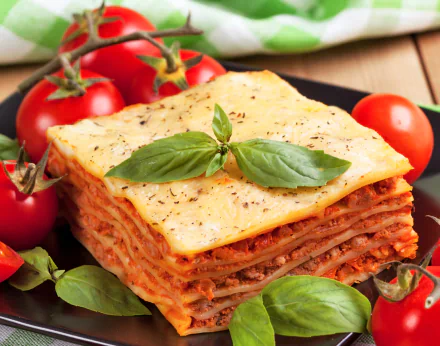 HD desktop wallpaper featuring a close-up of a layered lasagna topped with fresh basil, surrounded by ripe tomatoes, highlighting rich colors and textures of the food.
