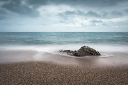 HD PC desktop wallpaper showing a tranquil ocean horizon over a sandy beach with a lone rock, soft waves and a cloudy sky — a nature background.