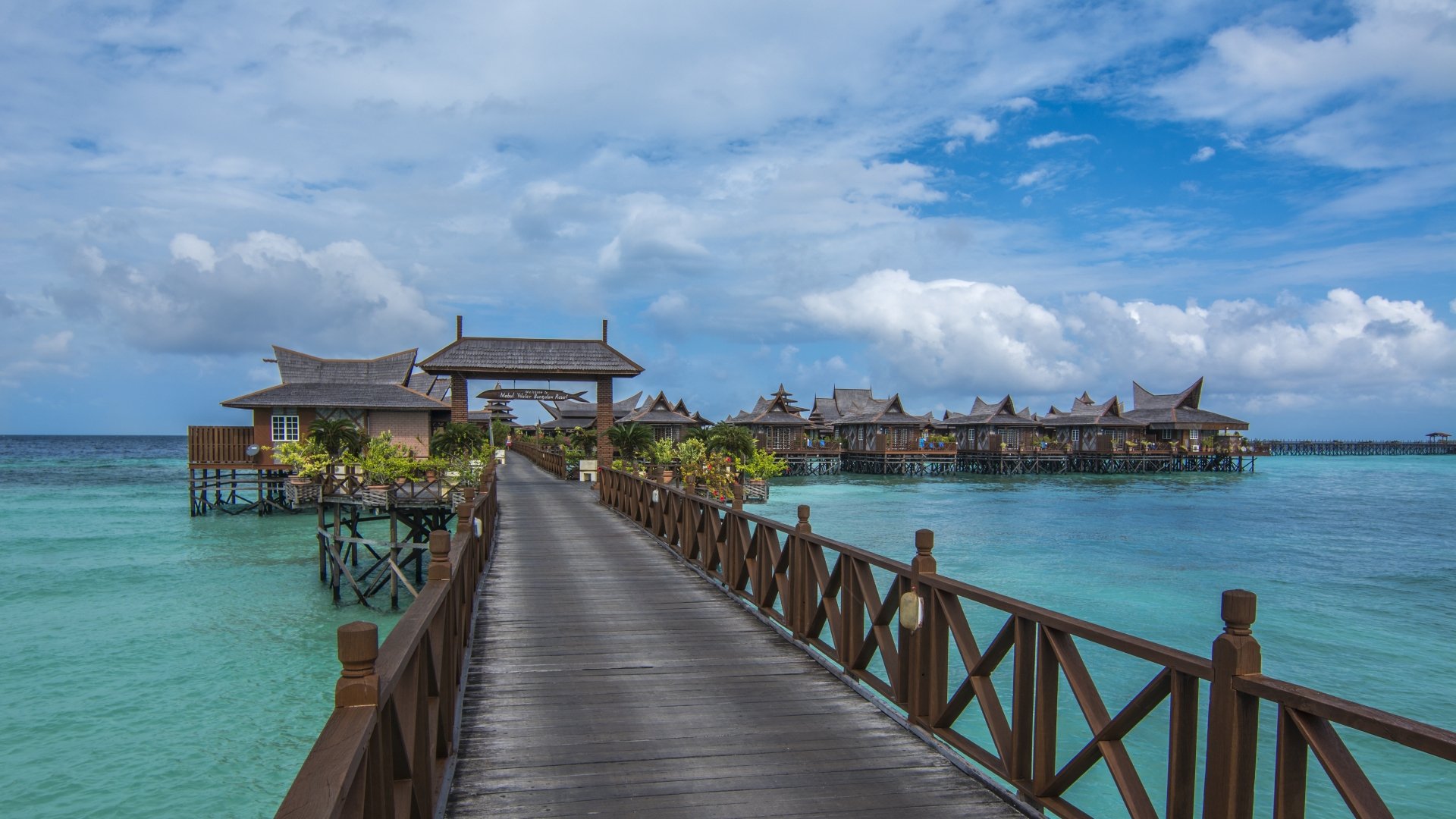 A wooden pier extends over the clear blue sea to tropical Malaysian bungalows set against a bright sky in this 4K Ultra HD ocean desktop wallpaper.