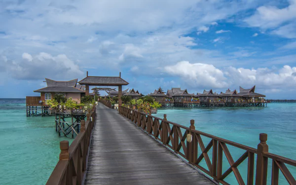 A wooden pier extends over the clear blue sea to tropical Malaysian bungalows set against a bright sky in this 4K Ultra HD ocean desktop wallpaper.
