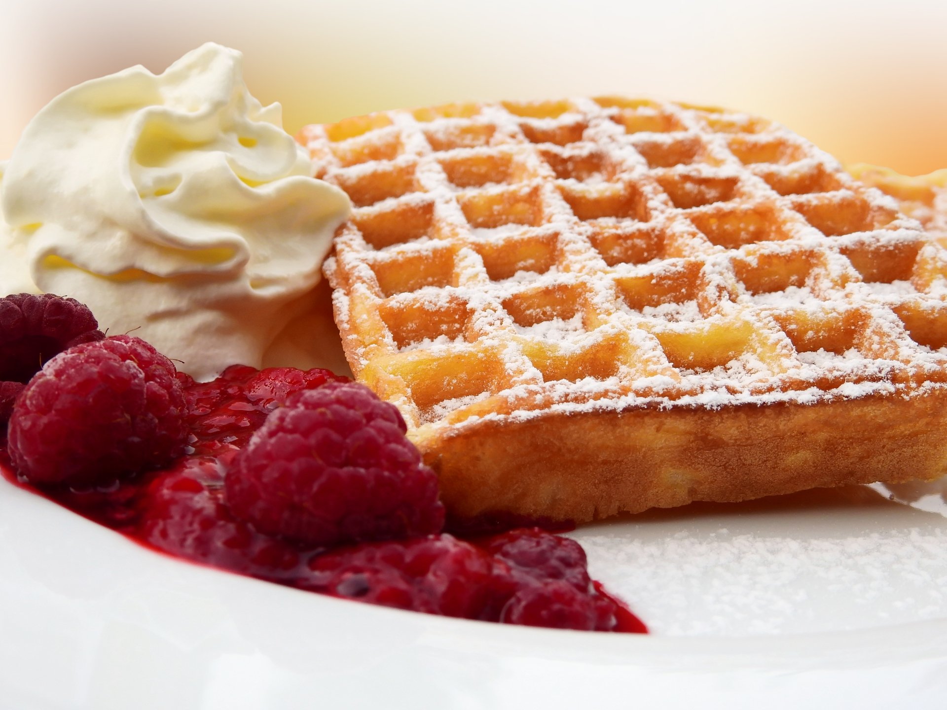 A close-up of a golden waffle dusted with powdered sugar, served with fresh raspberries and a dollop of cream, captured in 4K Ultra HD for a vibrant breakfast wallpaper.