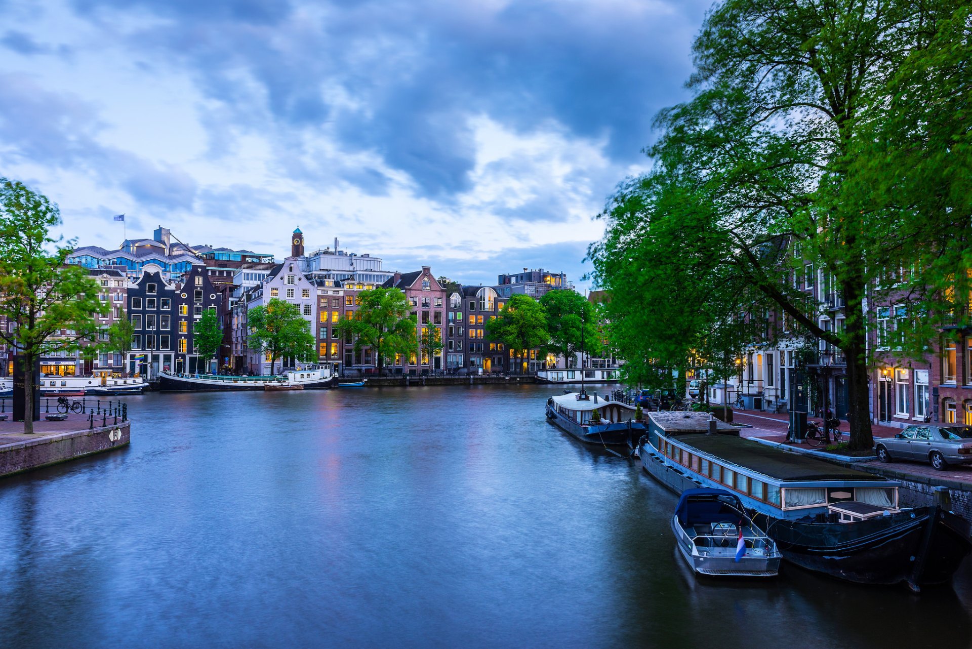 HD desktop wallpaper of an Amsterdam canal with tree-lined banks, historic houses, moored boats and man-made quays in a Netherlands city.