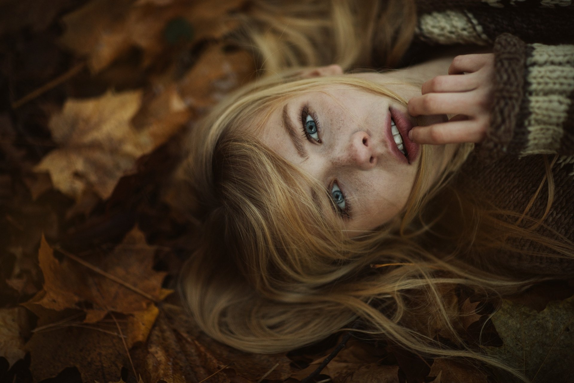 A blonde woman with blue eyes lies among autumn leaves, gazing softly at the camera, her finger delicately touching her lips, creating an intimate and serene atmosphere.