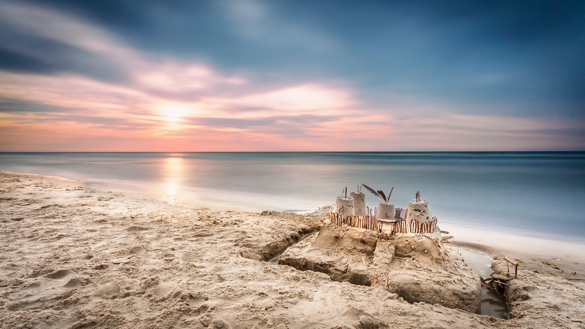 HD PC desktop wallpaper of a sand castle on the beach at sunrise, soft pastel sky, calm ocean meeting the horizon, and smooth sand in a serene nature scene.
