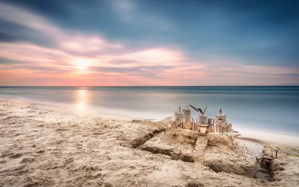 HD PC desktop wallpaper of a sand castle on the beach at sunrise, soft pastel sky, calm ocean meeting the horizon, and smooth sand in a serene nature scene.