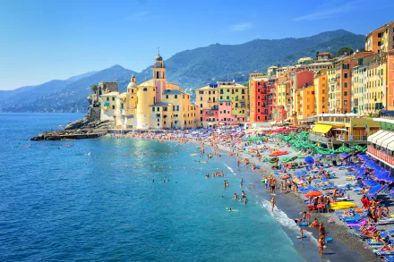 A vibrant beach scene in Genoa, Italy, featuring colorful houses lining the shore and people enjoying the sun and sea. The mountainous backdrop enhances the picturesque setting.