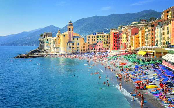 A vibrant beach scene in Genoa, Italy, featuring colorful houses lining the shore and people enjoying the sun and sea. The mountainous backdrop enhances the picturesque setting.