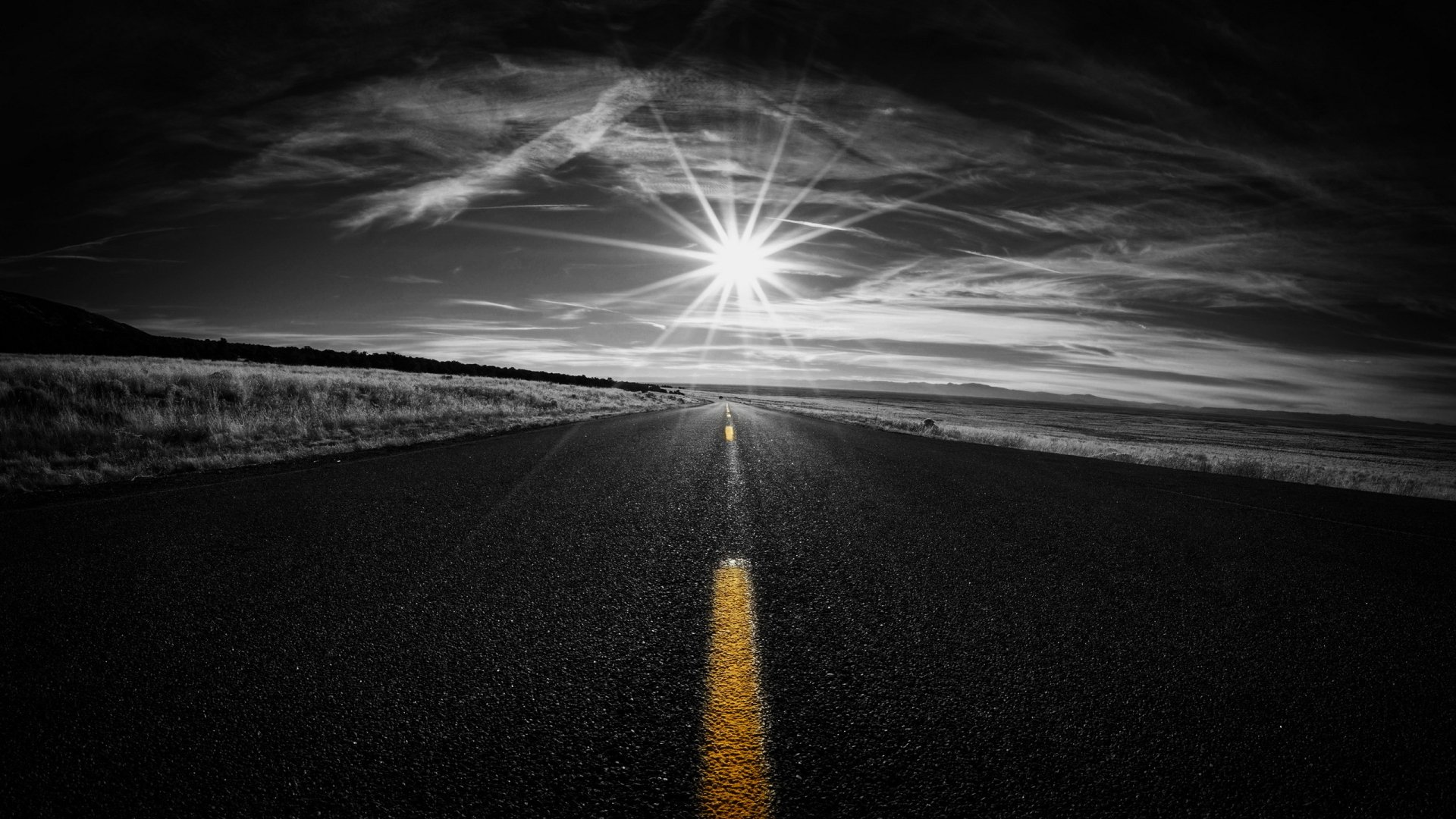 Black & white HD wallpaper of a road stretching to the horizon under a sunbeam-filled sky, featuring selective color on the yellow road line.