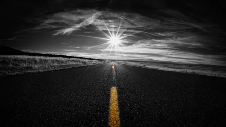 Black & white HD wallpaper of a road stretching to the horizon under a sunbeam-filled sky, featuring selective color on the yellow road line.