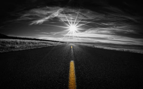 Black & white HD wallpaper of a road stretching to the horizon under a sunbeam-filled sky, featuring selective color on the yellow road line.
