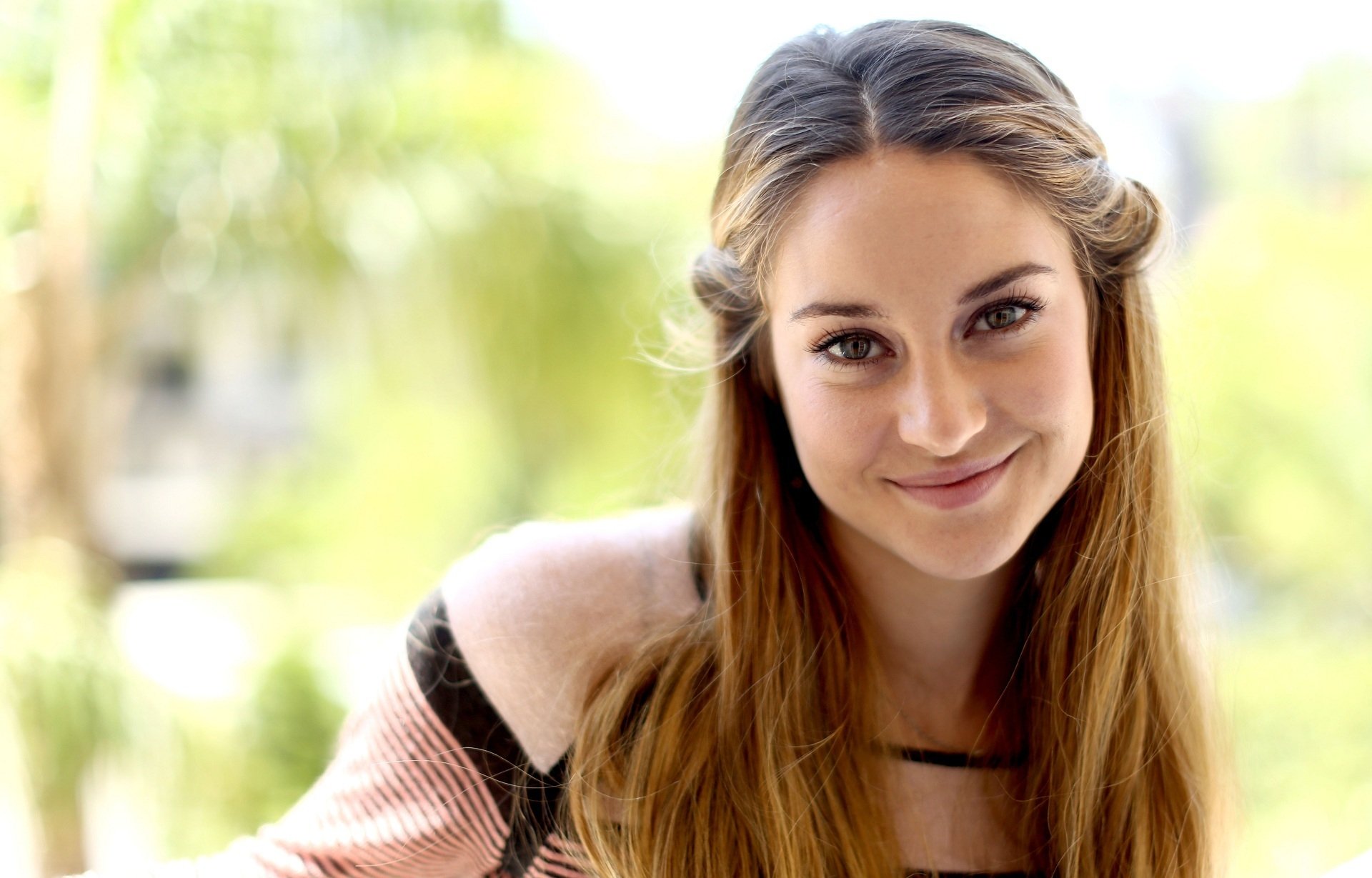 HD wallpaper featuring actress Shailene Woodley. The image showcases her smiling, with long brunette hair, set against a blurred outdoor background.