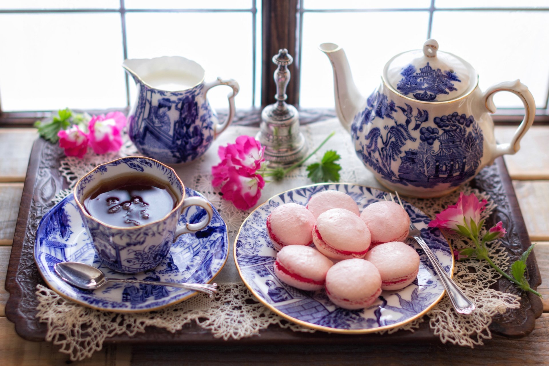 A 4K Ultra HD still life featuring a blue and white teapot, matching tea cup, pink macarons, and fresh flowers on a lace-covered wooden tray by a window.