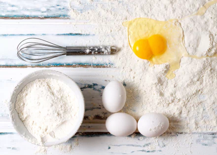HD PC desktop wallpaper of baking ingredients: bowl of flour, three eggs with a cracked double yolk, and a whisk on a flour-dusted rustic wooden surface.