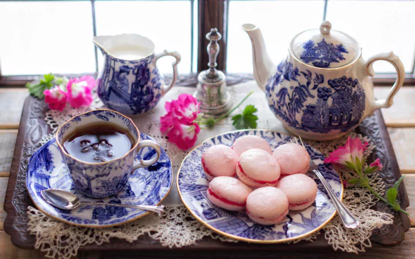 A 4K Ultra HD still life featuring a blue and white teapot, matching tea cup, pink macarons, and fresh flowers on a lace-covered wooden tray by a window.