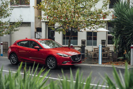 A red Mazda 3 compact car parked on a street with autumn trees and urban buildings, captured in a sharp 4K Ultra HD background scene.