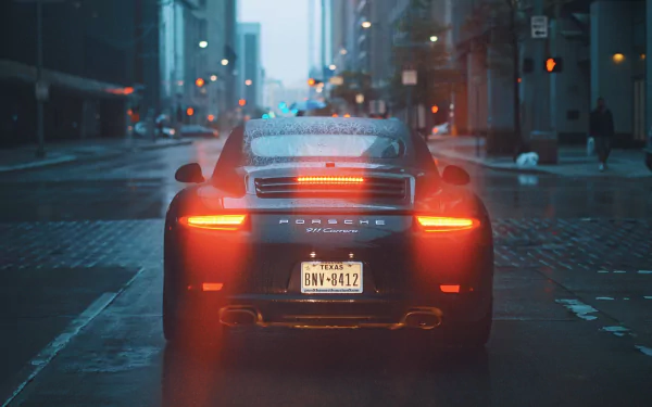 HD desktop wallpaper featuring a black Porsche 911 Carrera driving on a wet, rainy street lined with buildings and lit by streetlights.