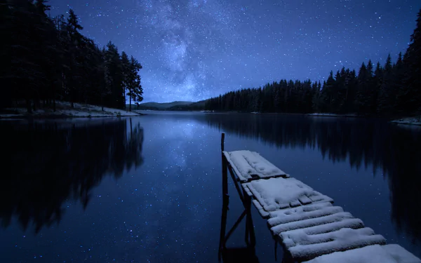 A snow-covered dock extends into a reflective lake under a starry winter sky, surrounded by trees. The serene scene captures the tranquil beauty of nature at night. HD desktop wallpaper.