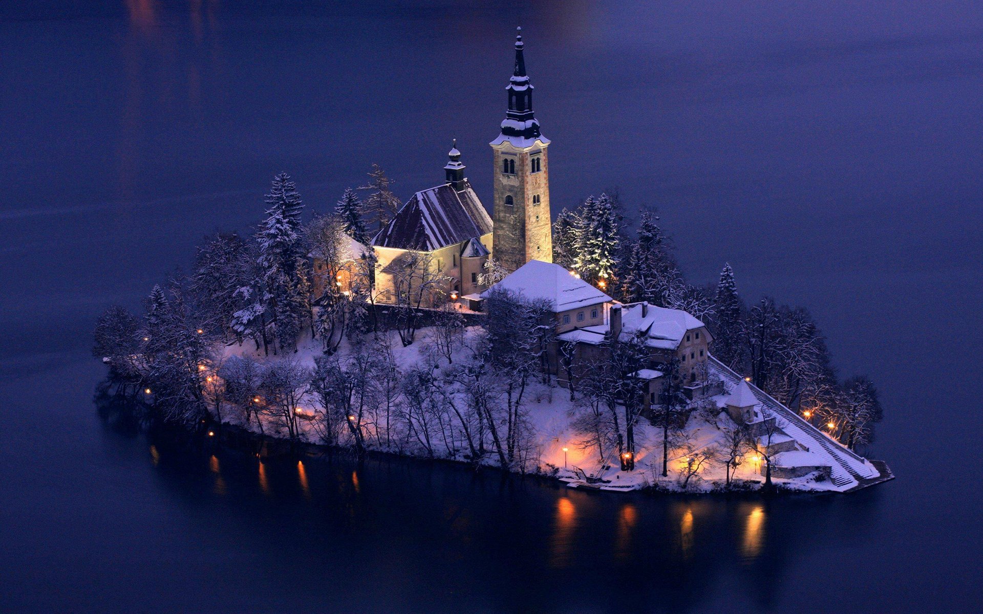 Snow-covered island with a church illuminated at night, surrounded by a calm lake in winter, captured in a high-definition photography wallpaper.