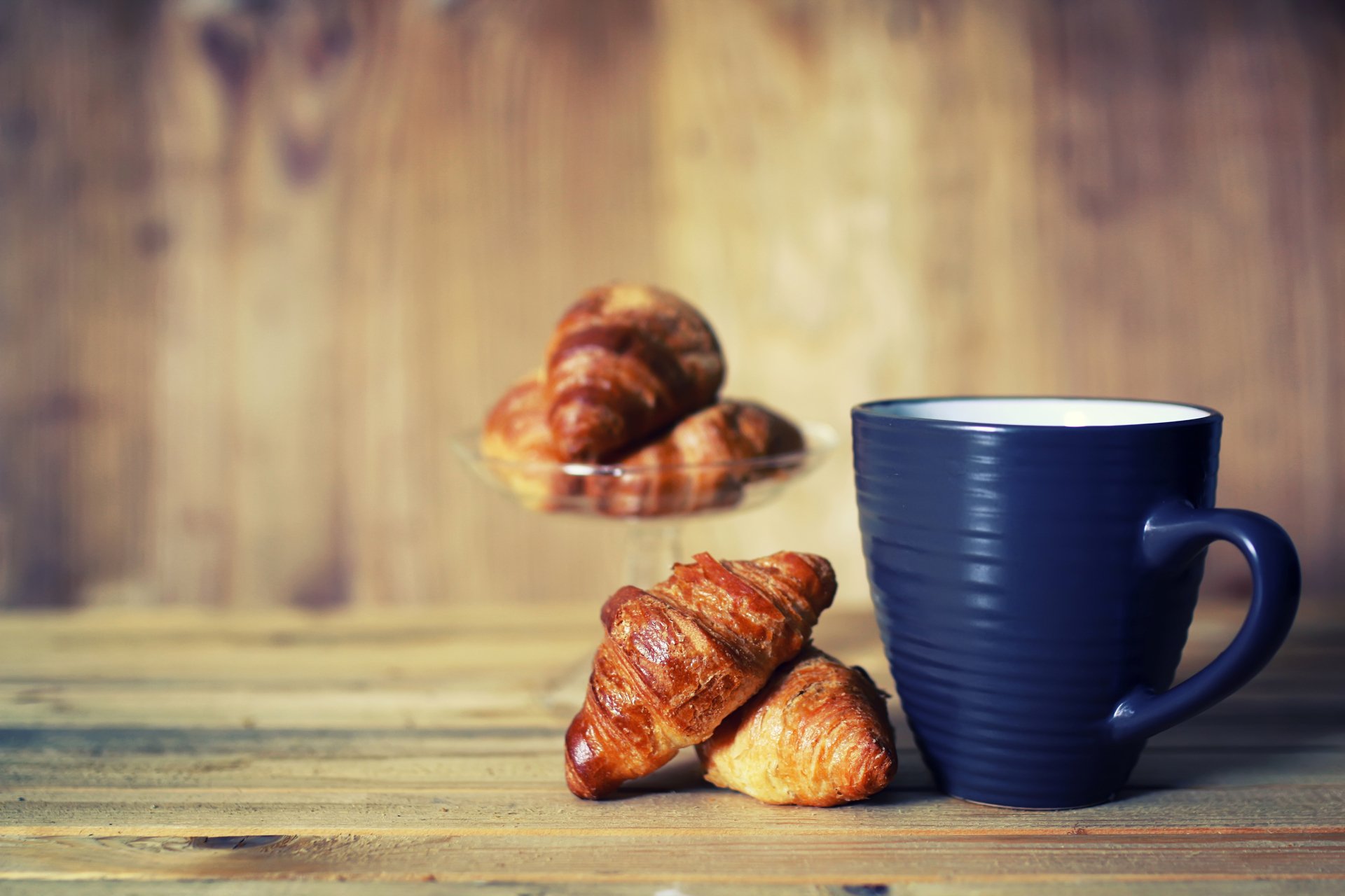 HD desktop wallpaper of a dark blue cup beside golden croissants on a wooden surface with a blurred rustic wooden background.