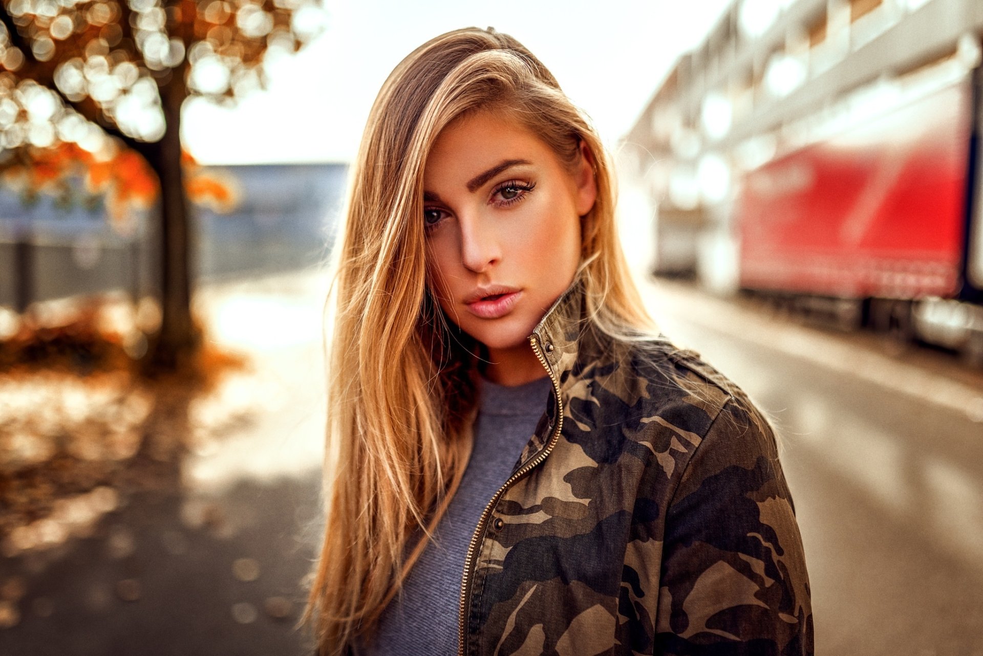 HD desktop wallpaper featuring a blonde woman model with brown eyes captured through a shallow depth of field on an urban street backdrop.