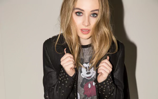 An HD desktop wallpaper featuring American singer Sabrina Carpenter with blonde hair and blue eyes, wearing a Mickey Mouse shirt and a studded black jacket.