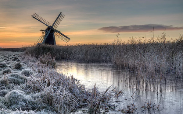 A frozen winter landscape featuring a man-made windmill beside icy plants and a reflective waterway, captured in stunning 4K Ultra HD quality.