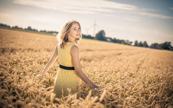 A blonde woman in a yellow dress stands in a wheat field during summer, with a blurred background enhancing the mood. The image is a high-definition desktop wallpaper and background.